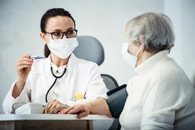 Ärztin spricht mit älterer Patientin, wobei beide eine Maske tragen
