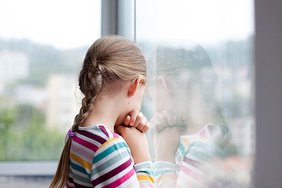 ein kleines Mädchen steht mit dem Rücken zum Betrachter und sieht traurig aus dem Fenster